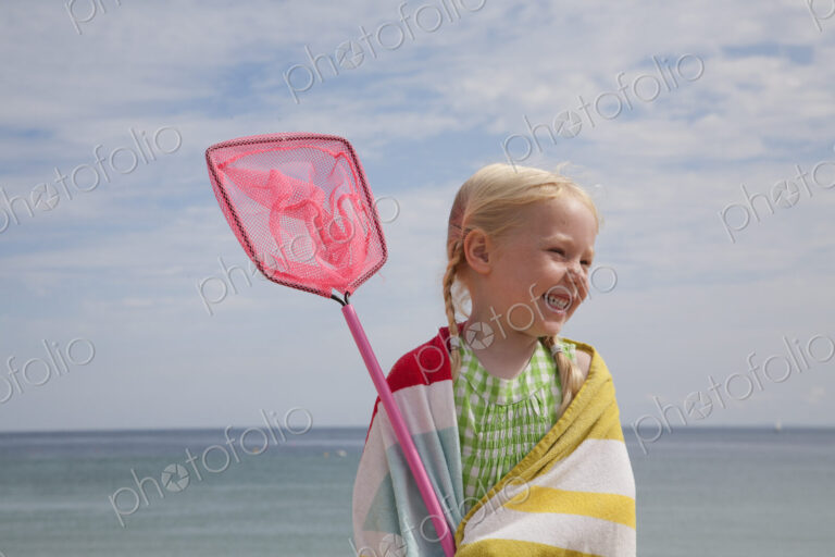 A young girl with a beach towel around her shoulders, carrying a small fishing net.