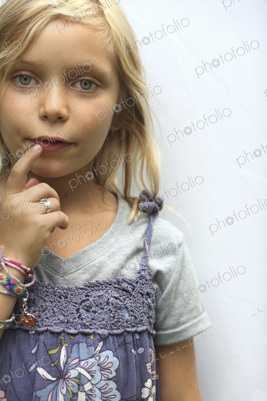 A young girl with blonde hair, and blue eyes. Her hand to her mouth.