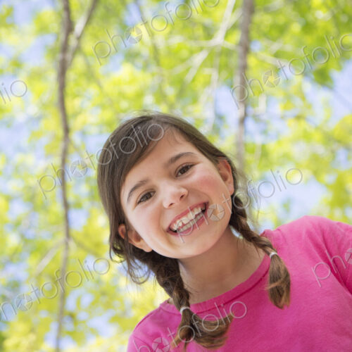 A young girl with braids wearing a pink top under a canopy of trees.