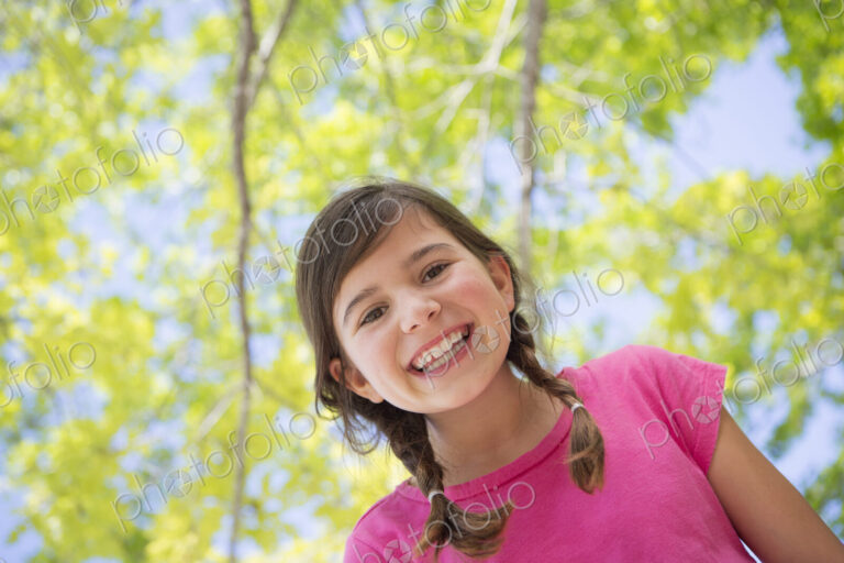 A young girl with braids wearing a pink top under a canopy of trees.