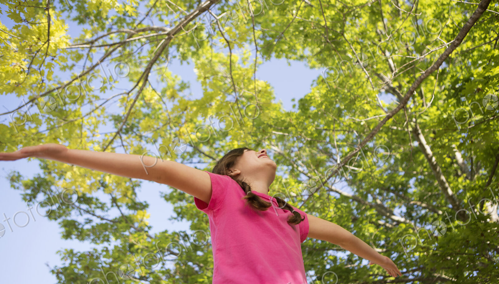 A young girl with braids wearing a pink top with her arms outstretched and head back.