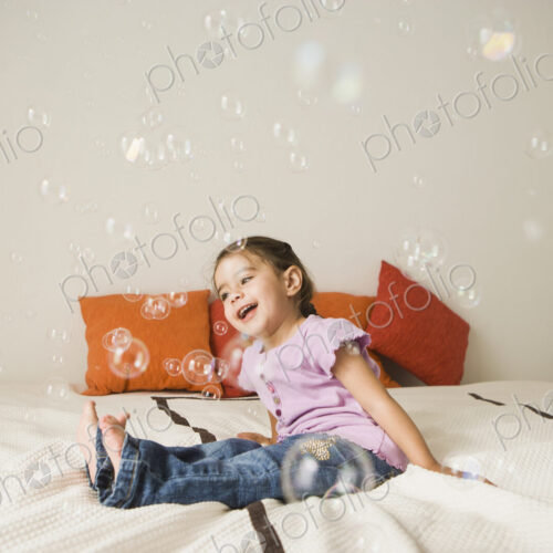 A young girl with brown eyes and dark hair in bunches sitting on a bed laughing.… A young girl with brown eyes and dark hair in bunches sitting on a bed laughing.…