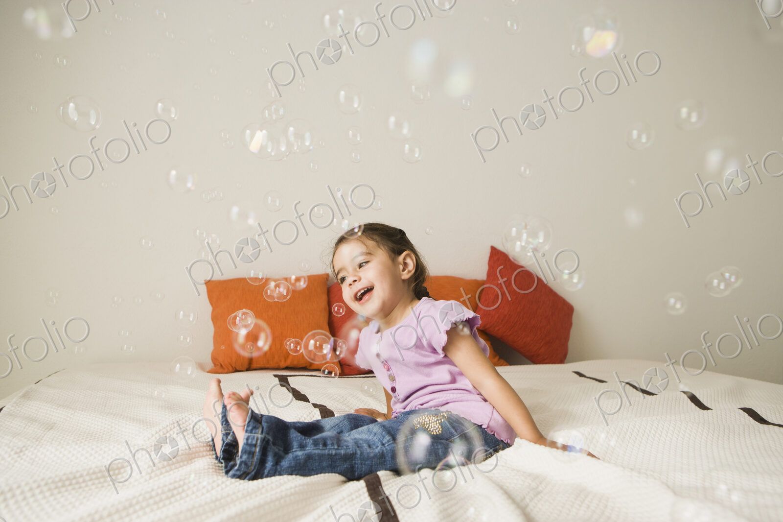 A young girl with brown eyes and dark hair in bunches sitting on a bed laughing.…