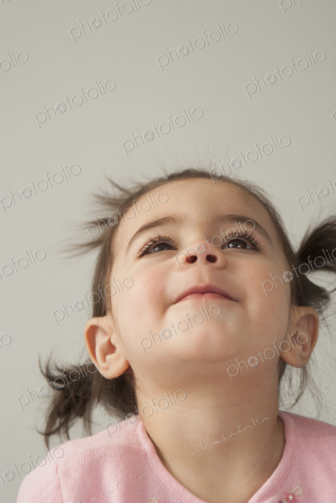 A young girl with brown eyes and dark hair in bunches. Looking upwards with her head…