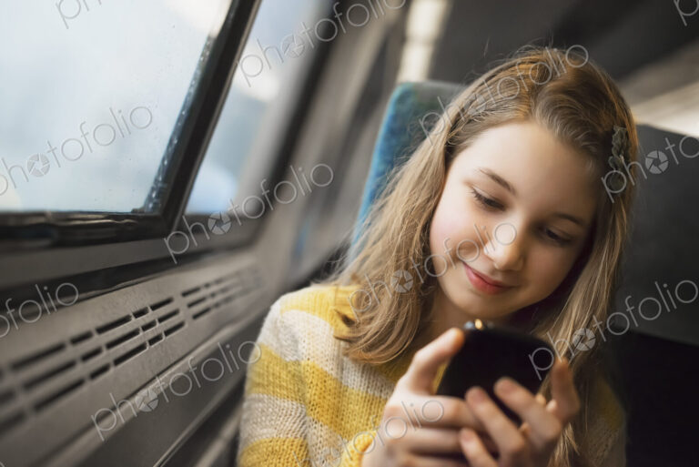 A young girl with long blonde hair sitting by a window on a train, using her mobile…