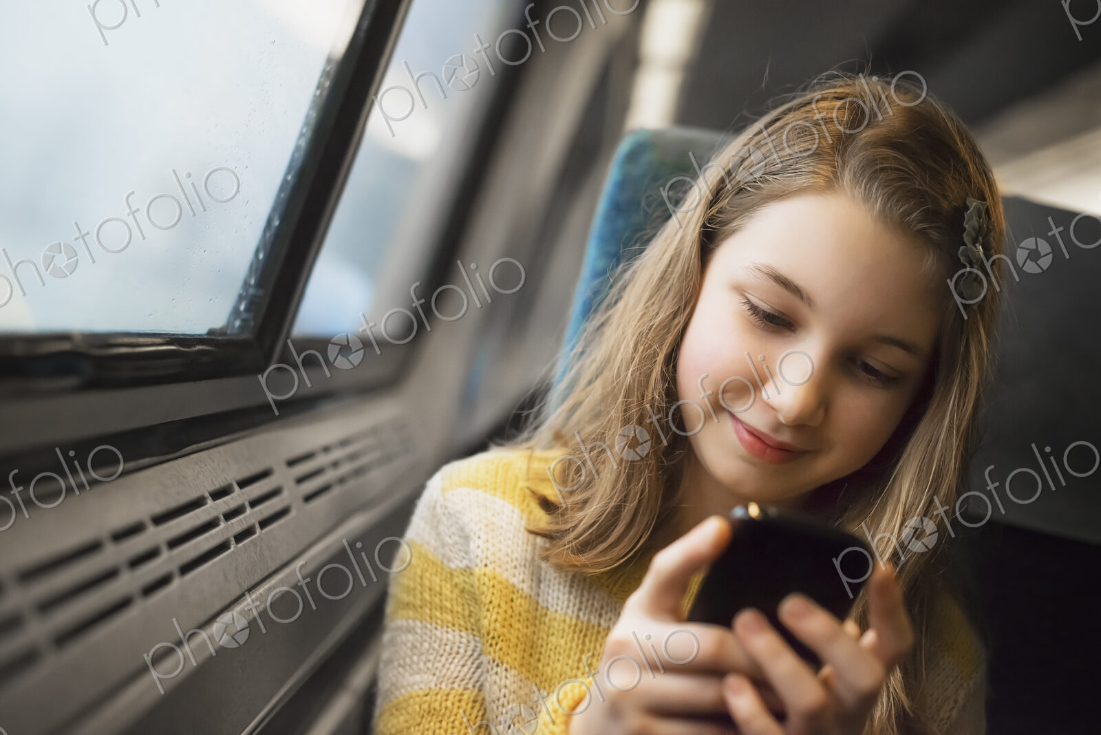 A young girl with long blonde hair sitting by a window on a train, using her mobile…