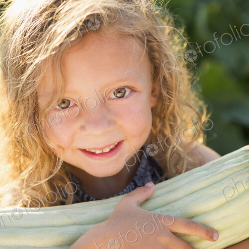 A young girl with long red curly hair outdoors in a garden holding a large fresh corn…