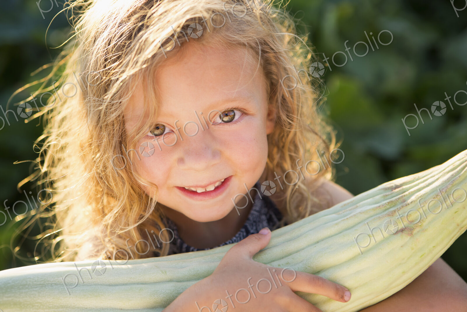 A young girl with long red curly hair outdoors in a garden holding a large fresh corn…