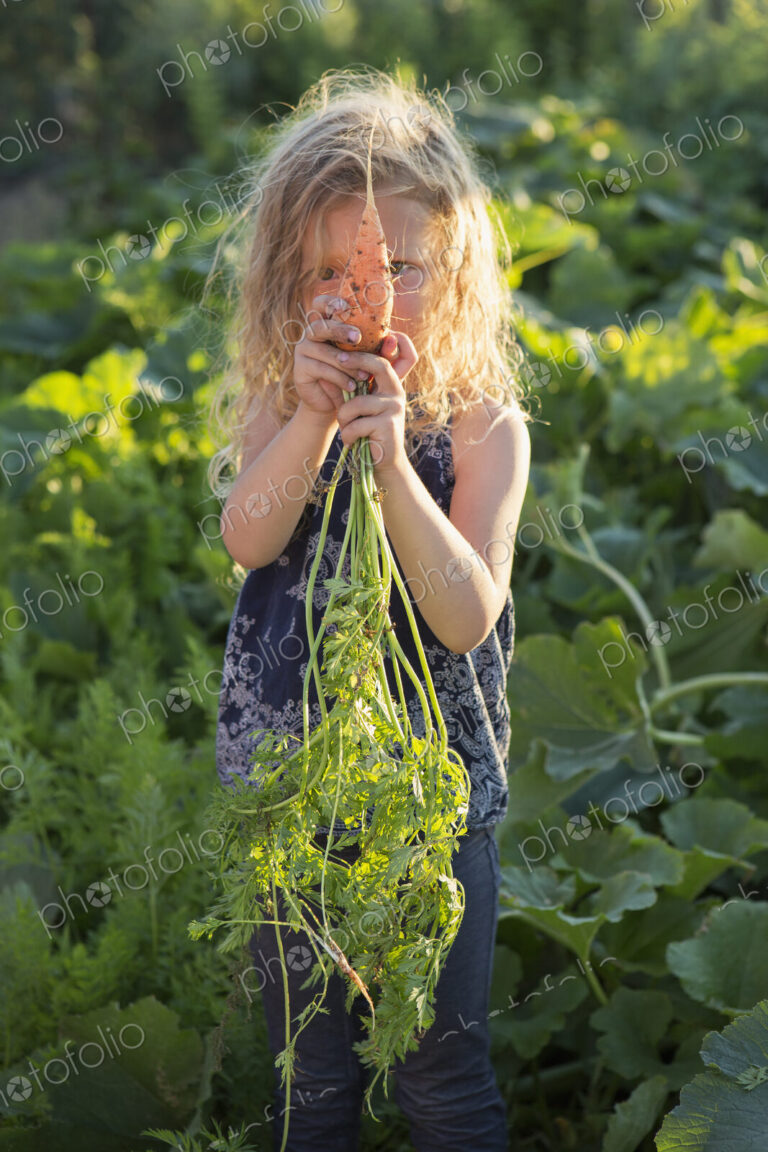 A young girl with long red curly hair outdoors in a garden, holding freshly picked…