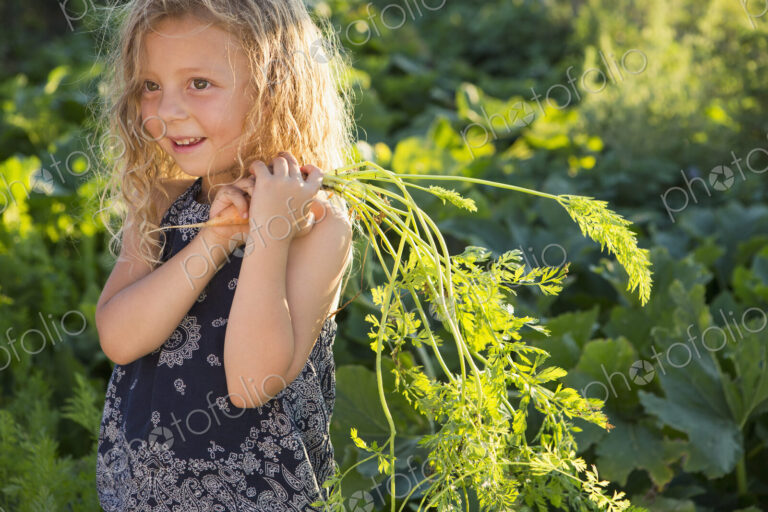 A young girl with long red curly hair outdoors in a garden, holding freshly picked…