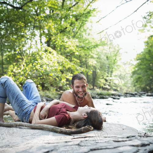 A young man and woman lying on the rocks on a river bank.