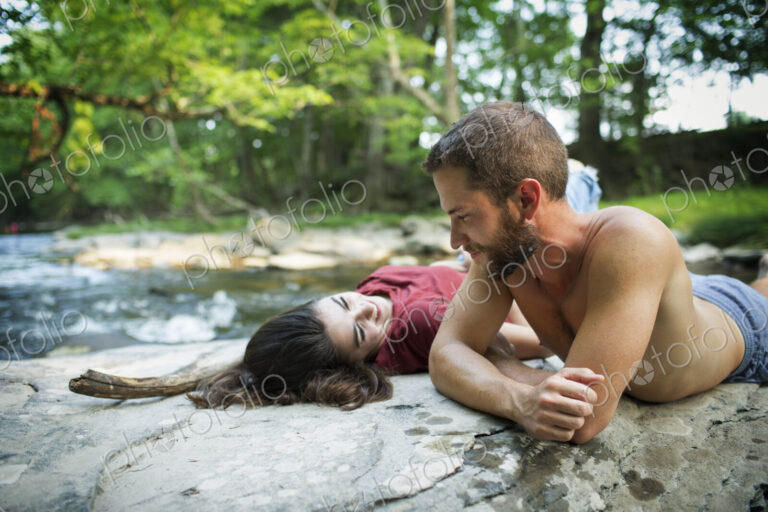A young man and woman lying on the rocks on a river bank.