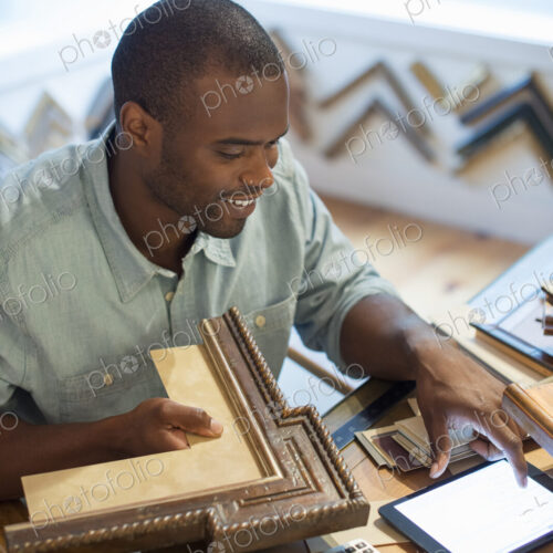 A young man at his workbench in a picture framing studio. Surrounded by samples. Using…