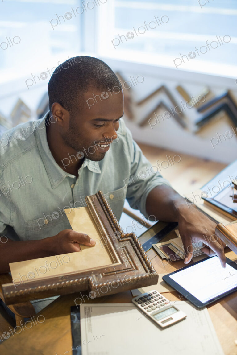 A young man at his workbench in a picture framing studio. Surrounded by samples. Using…