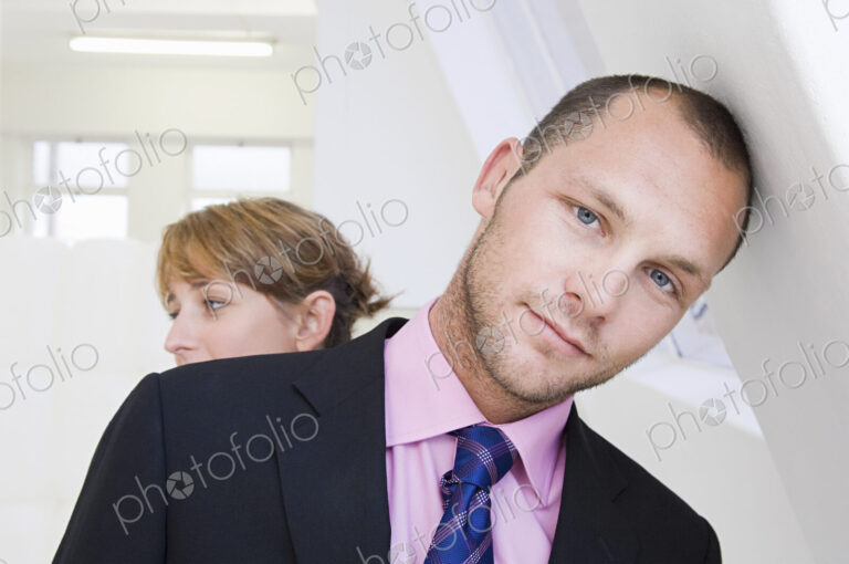 A young man in a suit and pink shirt, leaning against a wall, and a young woman in the…