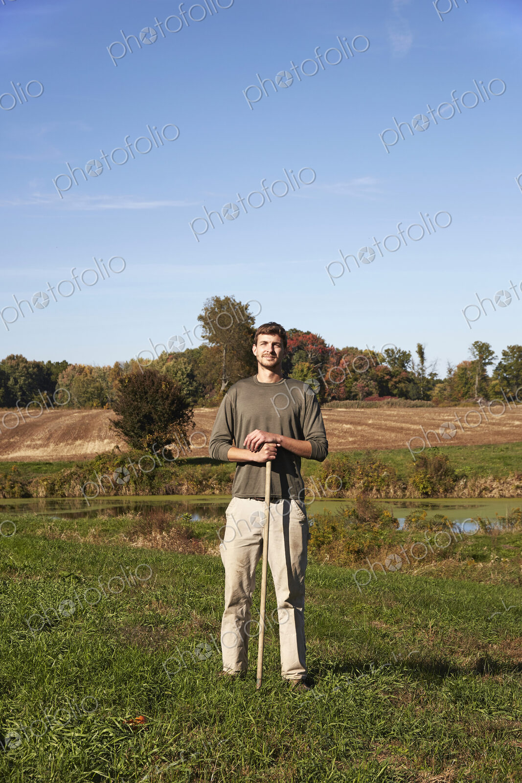 A young man in working clothes standing in a field, holding a tool.
