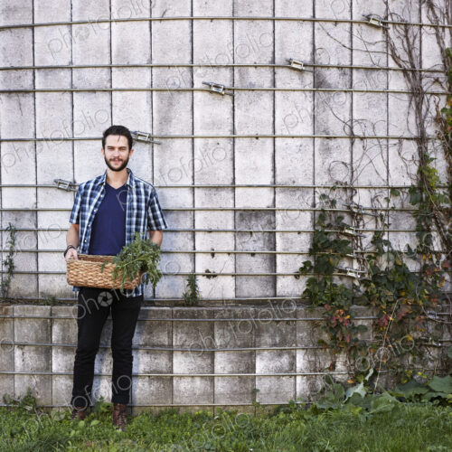 A young man in working clothes standing in front of a wall with plants growing up a… A young man in working clothes standing in front of a wall with plants growing up a…