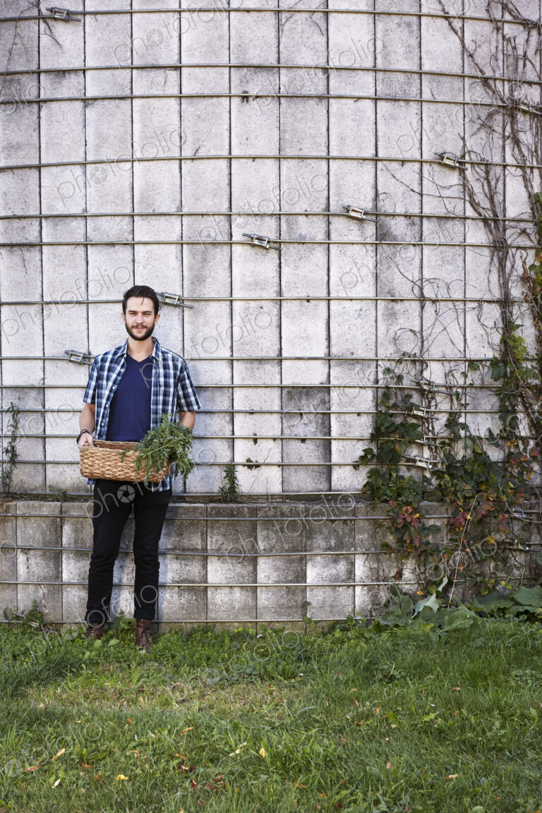 A young man in working clothes standing in front of a wall with plants growing up a…