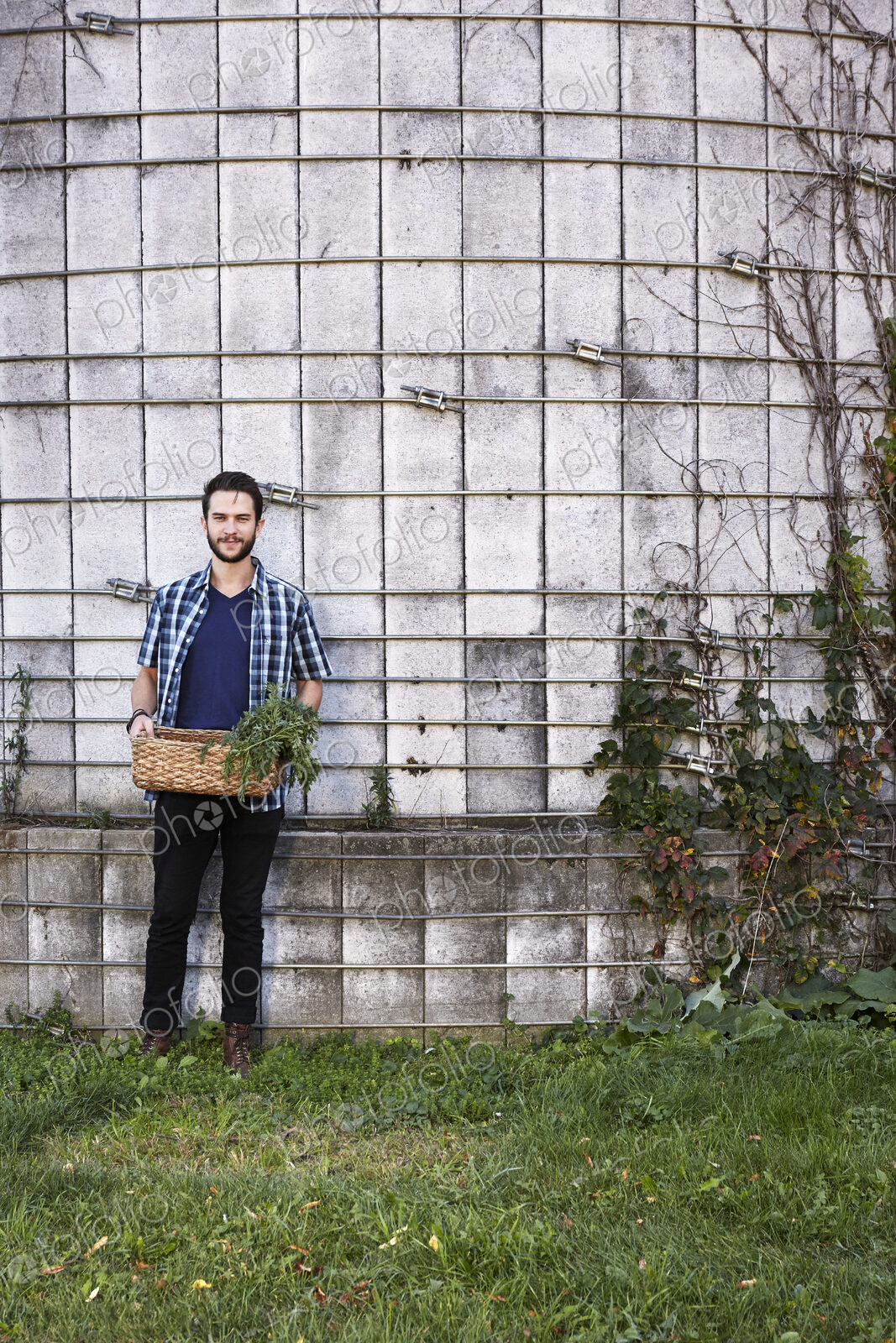 A young man in working clothes standing in front of a wall with plants growing up a…