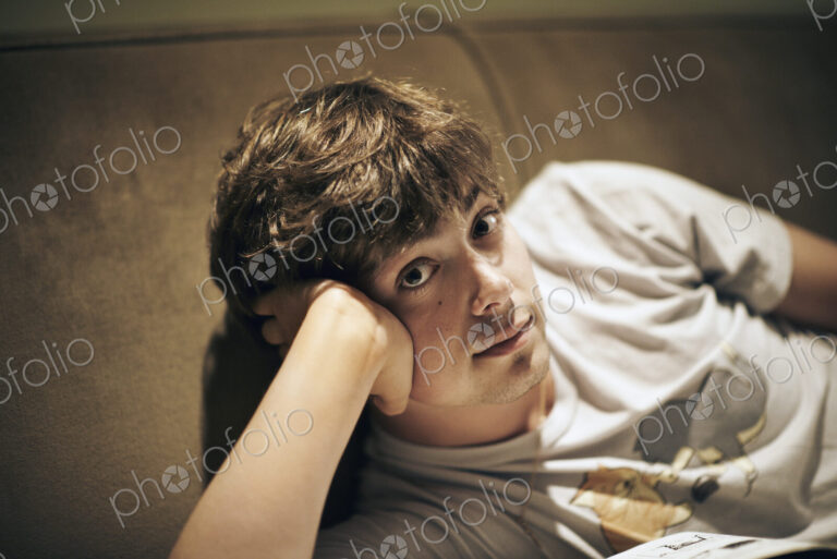 A young man lying on a sofa, resting his head on his elbow and looking at the camera.