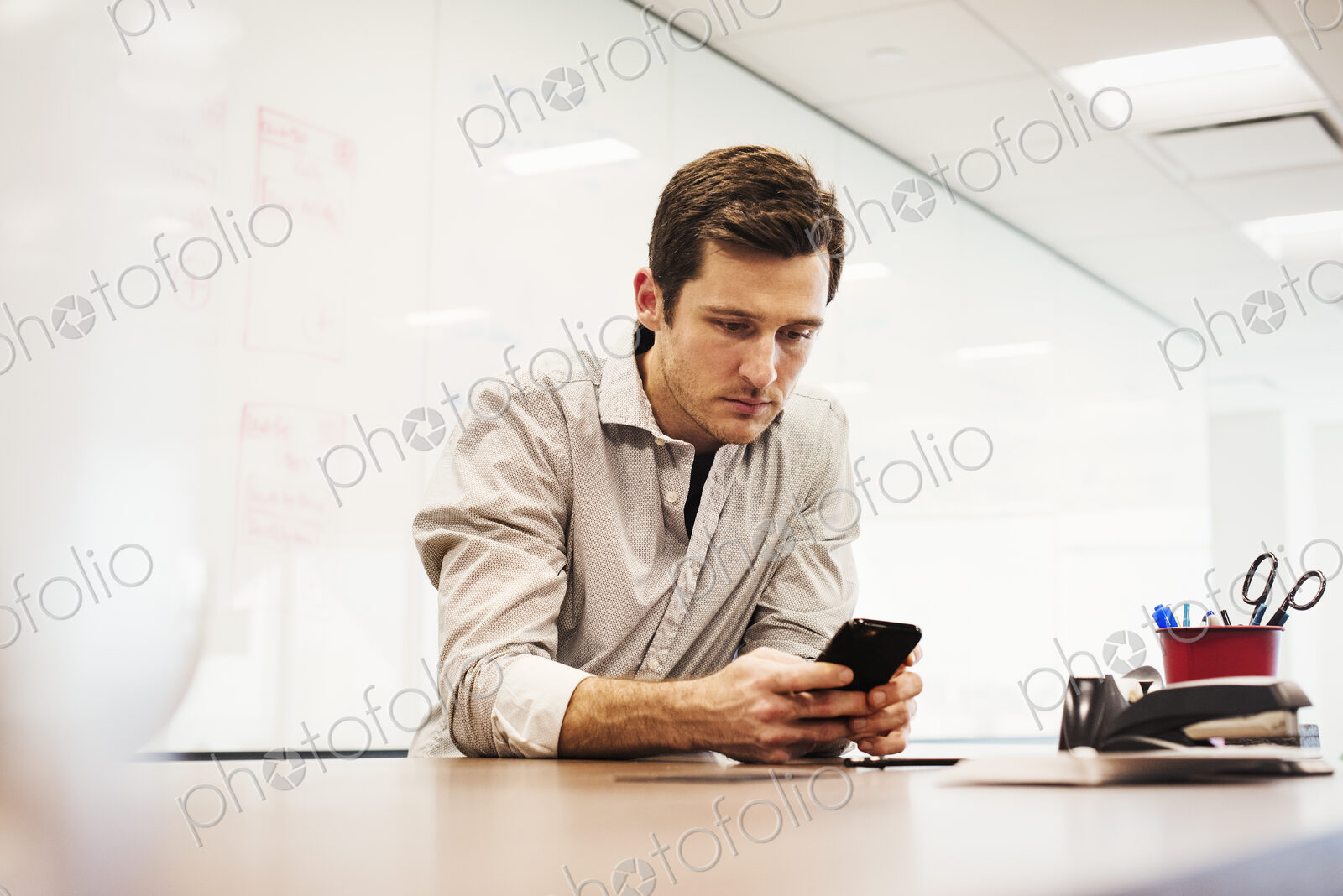 A young man sitting in a classroom at a table and looking down at a cellphone.