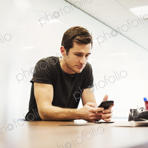 A young man sitting in a classroom leaning on a desk looking down at a cellphone. A young man sitting in a classroom leaning on a desk looking down at a cellphone.