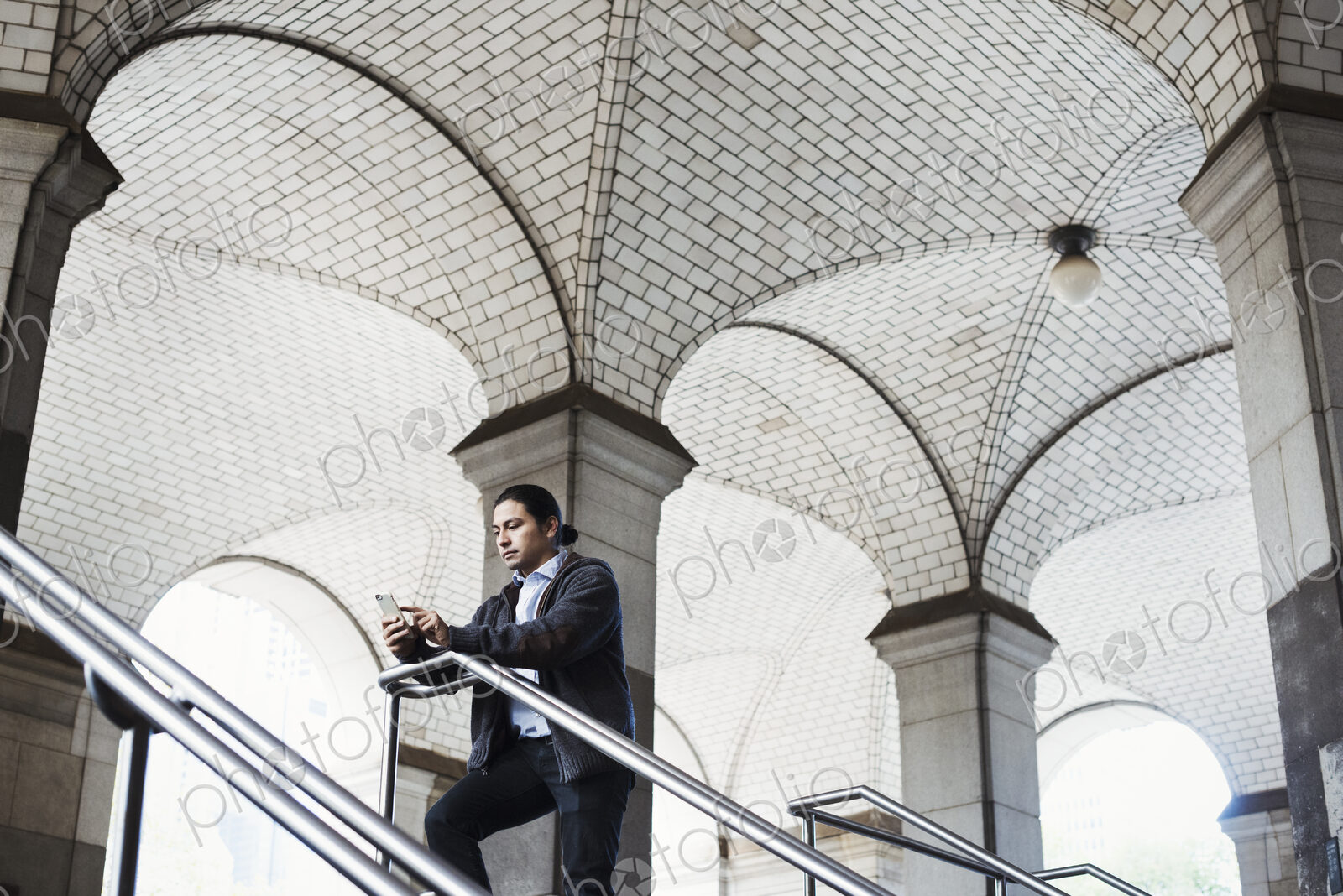 A young man standing on a stair under an archway, lookign at his mobile phone.