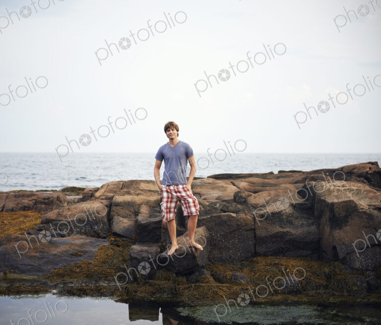 A young man standing on the shore by a rockpool with his hands in his pockets.