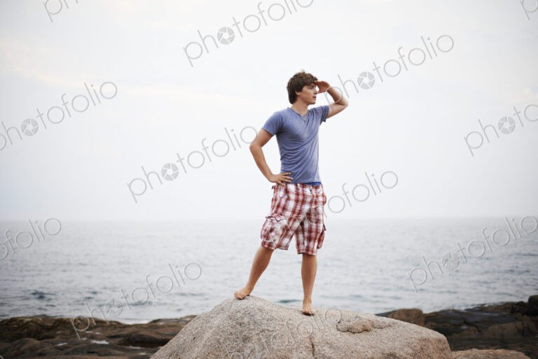 A young man with his hand to his head, shading his eyes and looking out from the…