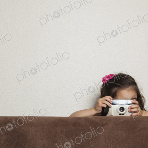 A young three year old girl crouching behind a sofa, with a toy camera, taking a picture.