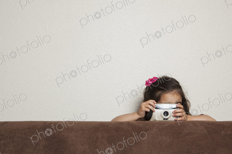 A young three year old girl crouching behind a sofa, with a toy camera, taking a picture.