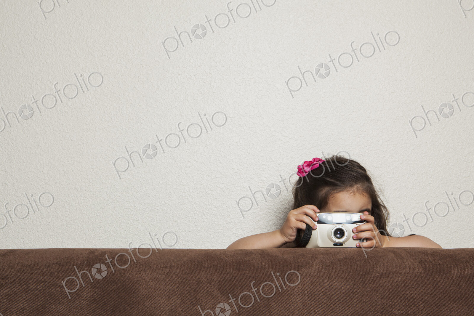 A young three year old girl crouching behind a sofa, with a toy camera, taking a picture.