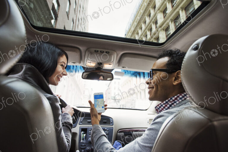 A young woman and young man in a car looking at a map on the display of a cellphone,…