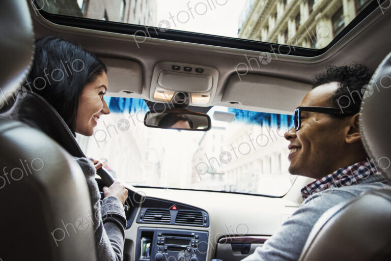 A young woman and young man in a car looking at each other and smiling, seen from the…