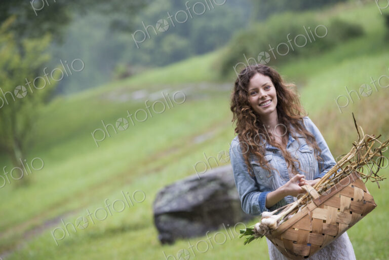 A young woman carrying a basket of freshly harvested garlic and vegetables.