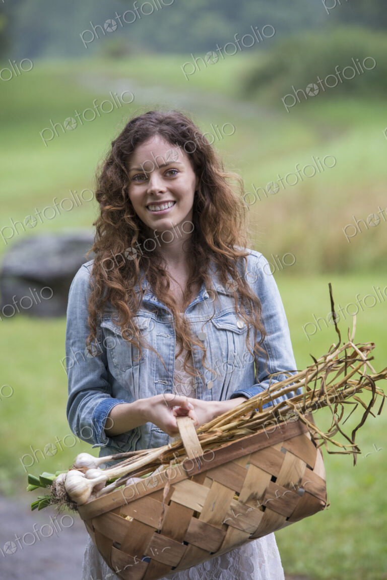 A young woman carrying a basket of freshly harvested garlic and vegetables.