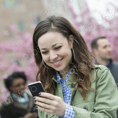 A young woman checking her smart phone for messages in the park. A group of friends in…