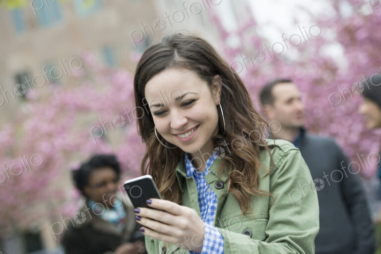 A young woman checking her smart phone for messages in the park. A group of friends in…