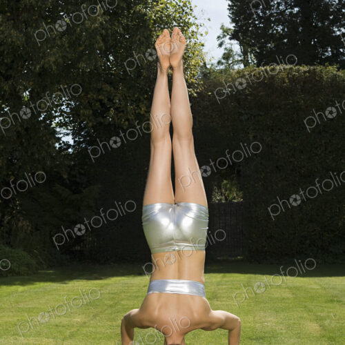 A young woman doing a handstand on grass, balancing on her head. Back view.