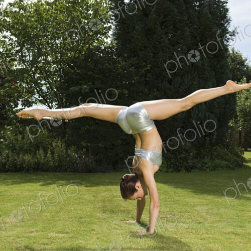 A young woman doing a splits handstand on the grass in a garden.
