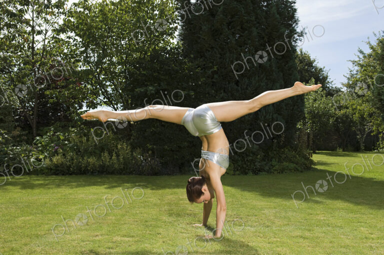 A young woman doing a splits handstand on the grass in a garden.