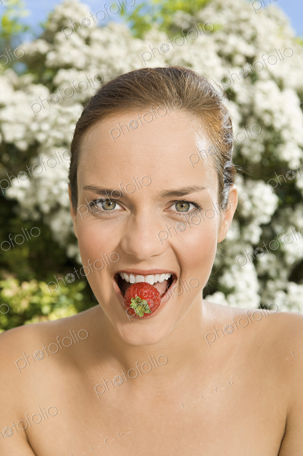 A young woman holding a strawberry between her teeth.