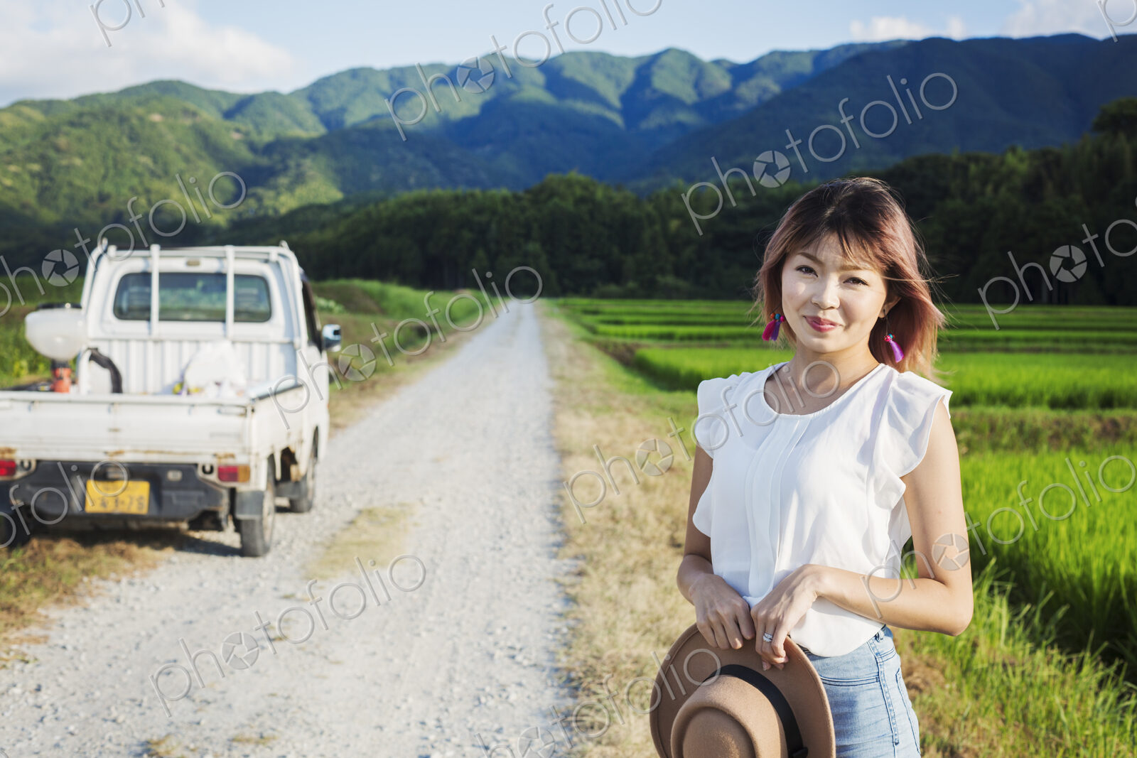 A young woman holding a sun hat, standing on a road by open fields of rice paddies.