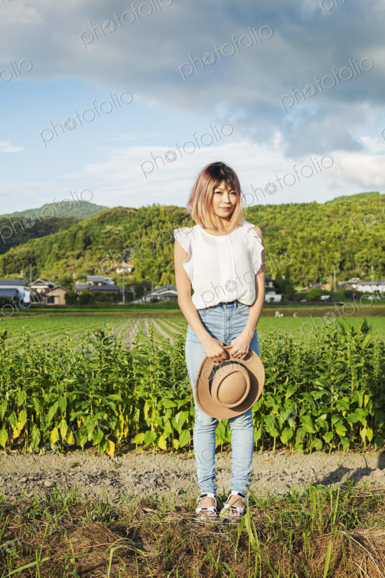 A young woman holding a sun hat, standing on a road by open fields of rice paddies.