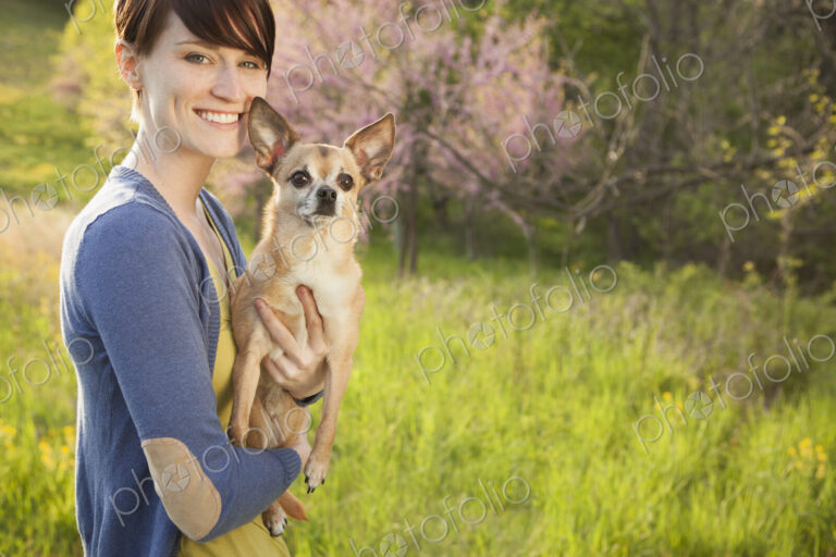 A young woman in a grassy field in spring. Holding a small chihuahua dog in her arms.…