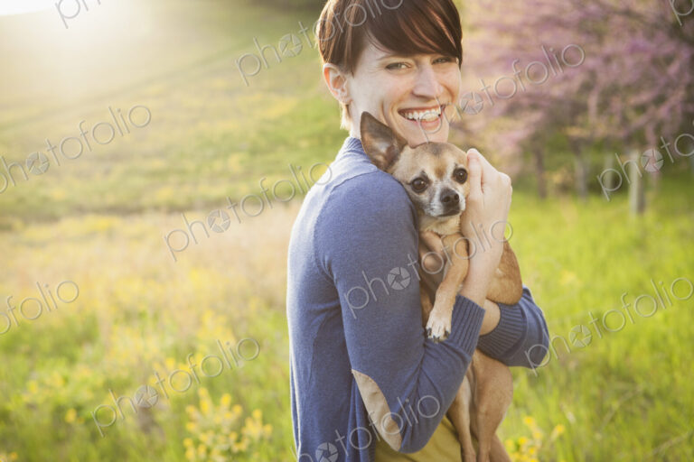 A young woman in a grassy field in spring. Holding a small chihuahua dog in her arms.…