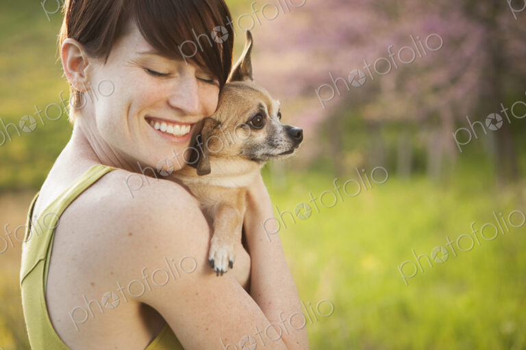 A young woman in a grassy field in spring. Holding a small chihuahua dog in her arms.…