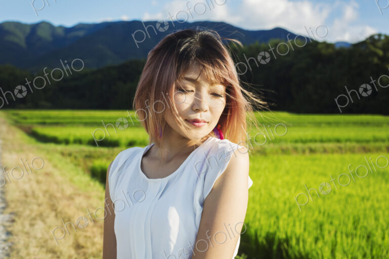 A young woman in a white shirt, hair blown in the wind, standing in open space by rice…