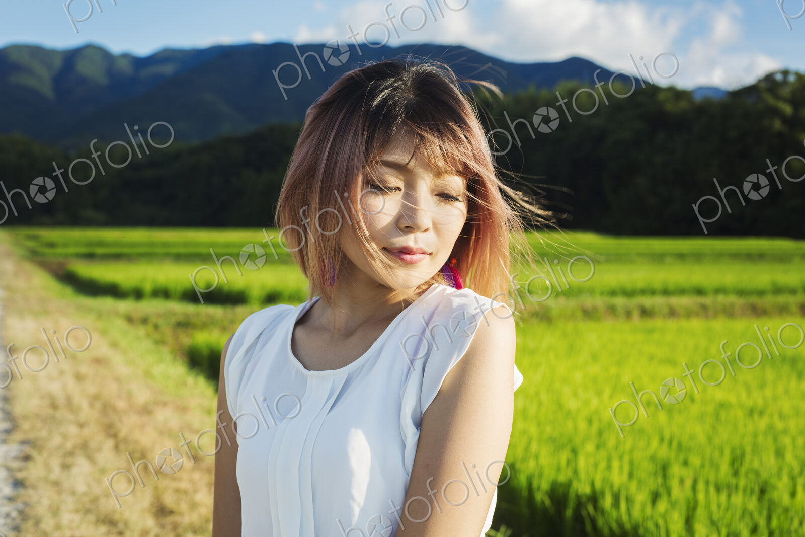 A young woman in a white shirt, hair blown in the wind, standing in open space by rice…