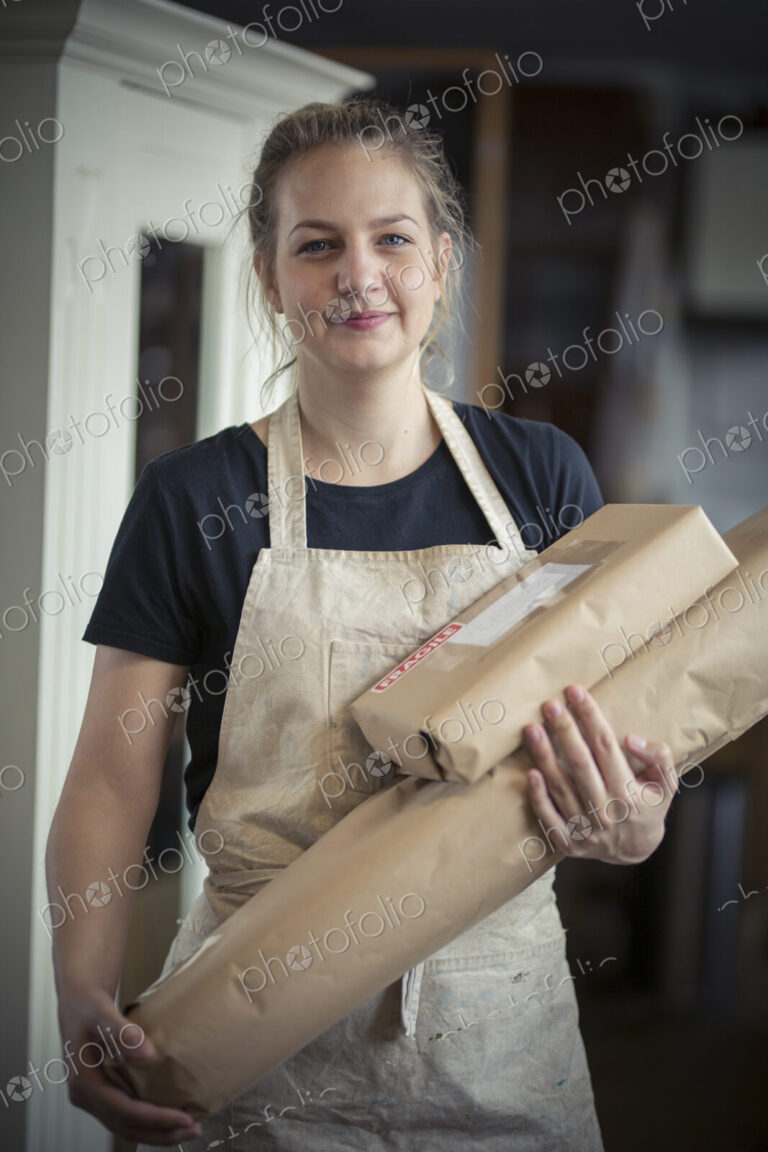A young woman in a workshop, holding two packages wrapped in brown packaging paper,…